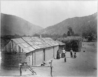 Indian family in front of adobe building and bough wigwam on reservation, Tejon Ranch, 1880.